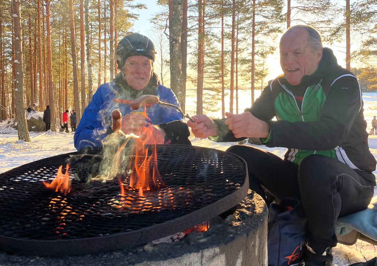 Twee schaatsers warmen zich op en roosteren eten bij een kampvuur tussen de dennen, naast het ijs