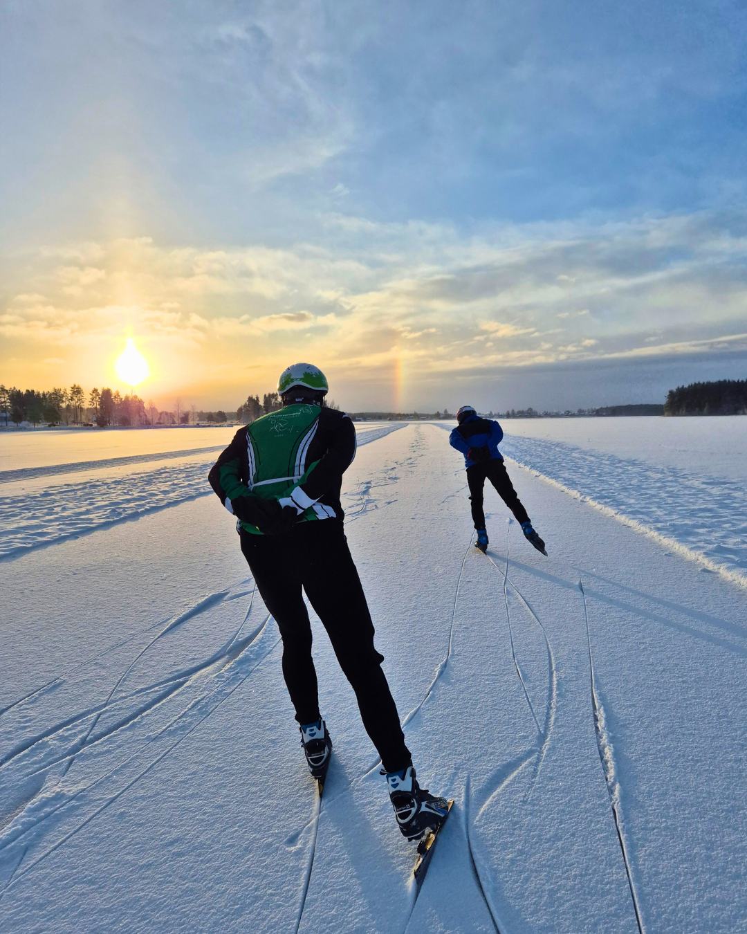 Twee schaatsers van achteren op een geveegde baan met een dun laagje sneeuw, richting de laagstaande zon